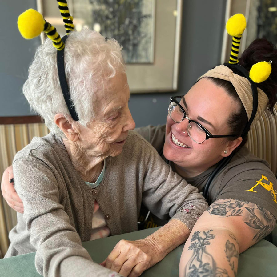 Senior resident and team member, both wearing bee antenna headbands, share a joyful, affectionate moment while smiling and embracing in a lively senior living community setting.