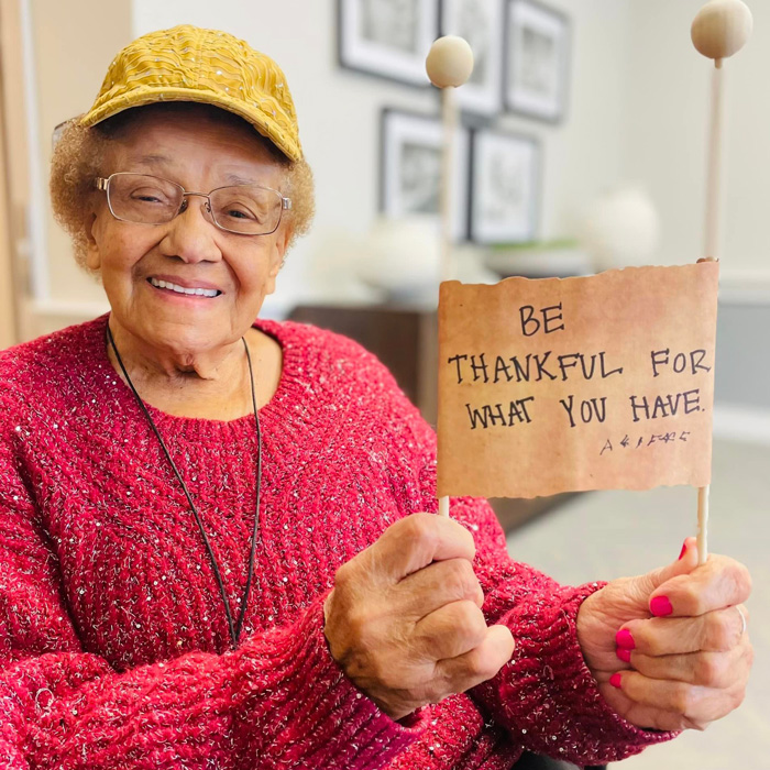 Senior resident smiling and holding a sign that reads 'Be Thankful for What You Have,' wearing a yellow hat and red sweater, promoting gratitude and positivity in community living.
