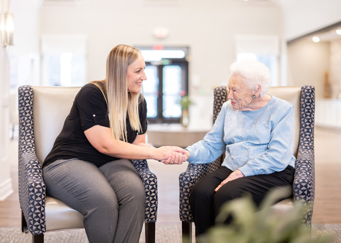 A smiling senior resident in a light blue sweater sits in a chair, holding hands with a friendly staff member in a senior living community, symbolizing care and connection.