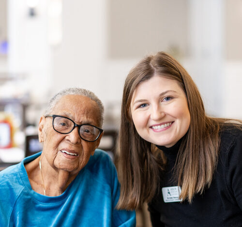 Senior resident in a blue sweater enjoys a cup of coffee while smiling alongside a friendly caregiver, emphasizing warmth, care, and companionship in the senior living community.