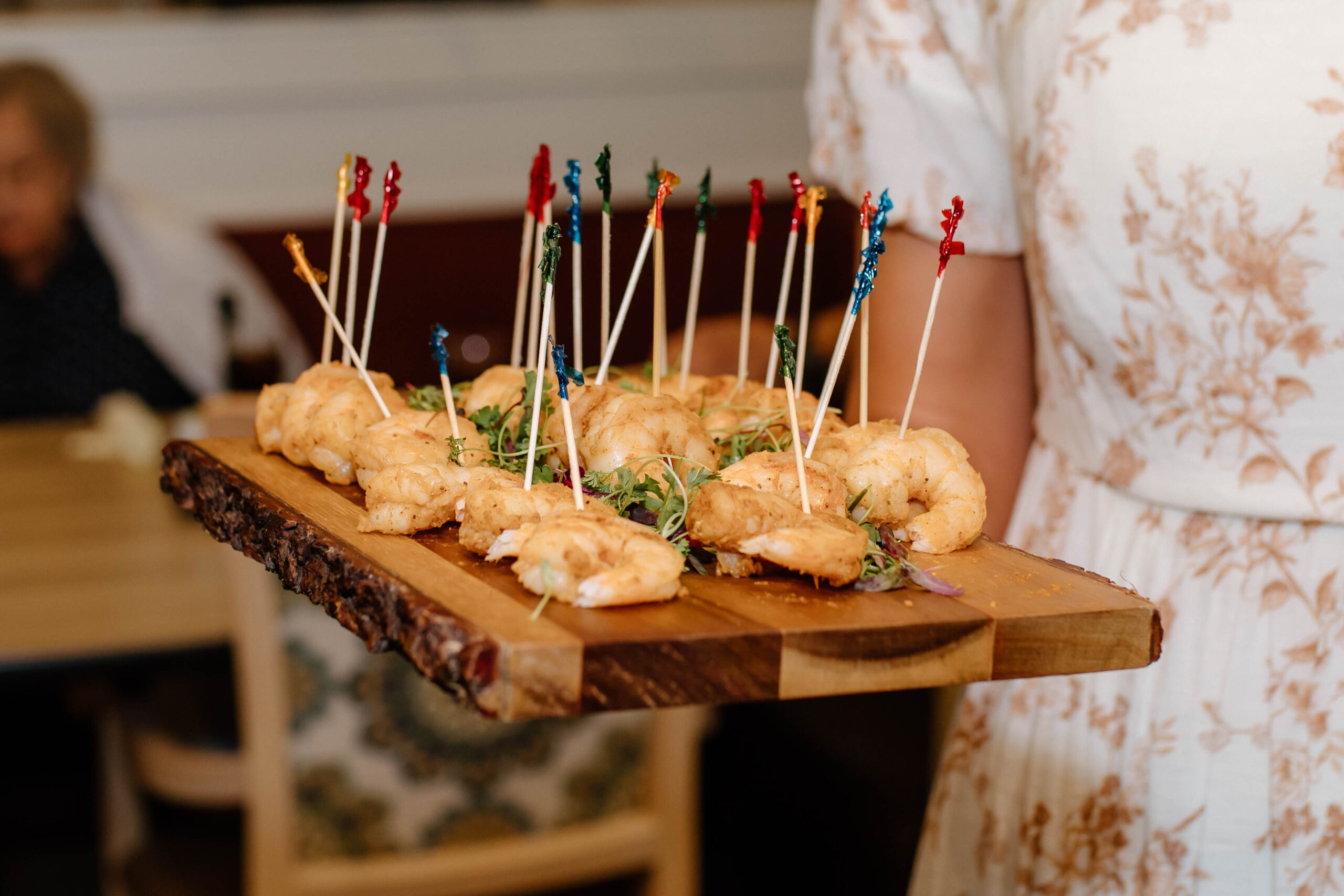 A server presenting a tray of gourmet shrimp tempura skewers, garnished with microgreens, on a polished wooden serving board in a dining room setting.