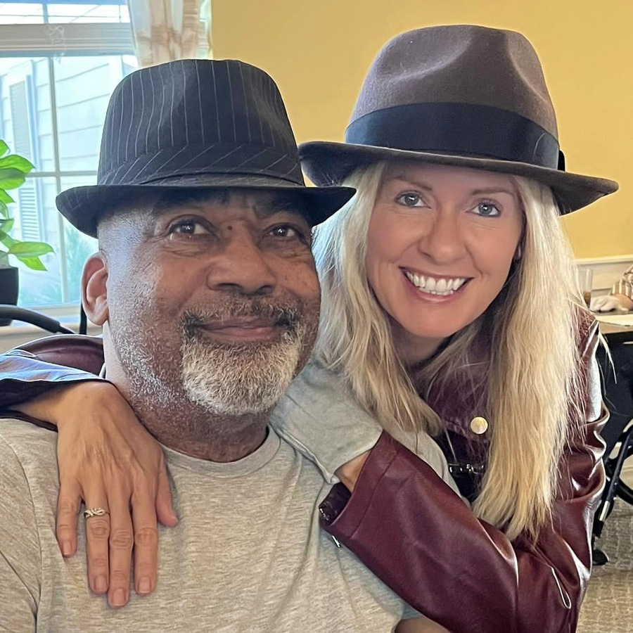 Two smiling individuals wearing stylish hats, one senior resident and one team member, embrace while enjoying a special event at the senior living community.