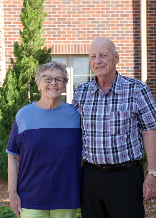 A senior resident couple stands outside in front of a brick building, smiling at the camera. The woman wears a blue and purple shirt, and the man wears a plaid shirt with black pants.