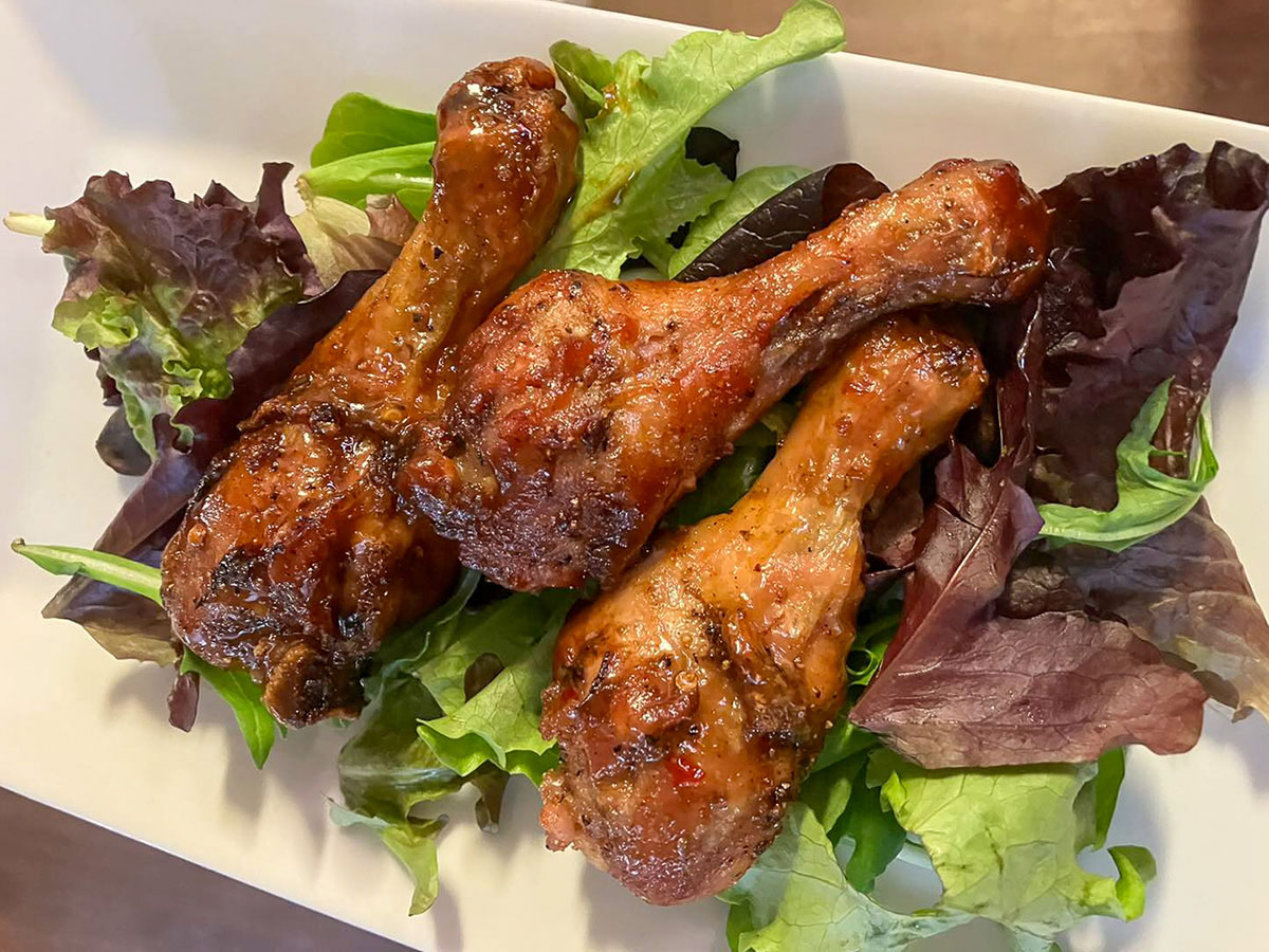 Three glazed chicken drumsticks served on a bed of fresh mixed greens, arranged on a white rectangular plate, showcasing a savory and appetizing meal presentation.