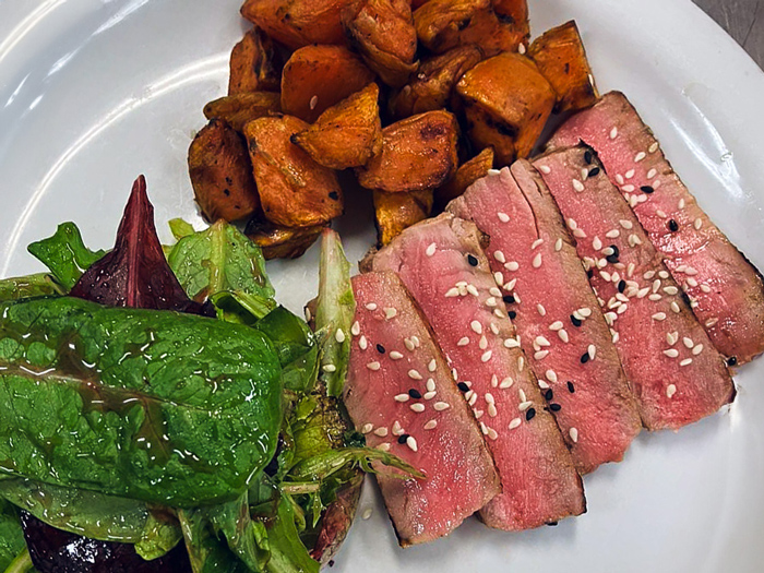 Plate featuring sesame-crusted seared tuna slices, roasted sweet potatoes, and a fresh mixed green salad, offering a colorful and balanced meal presentation.