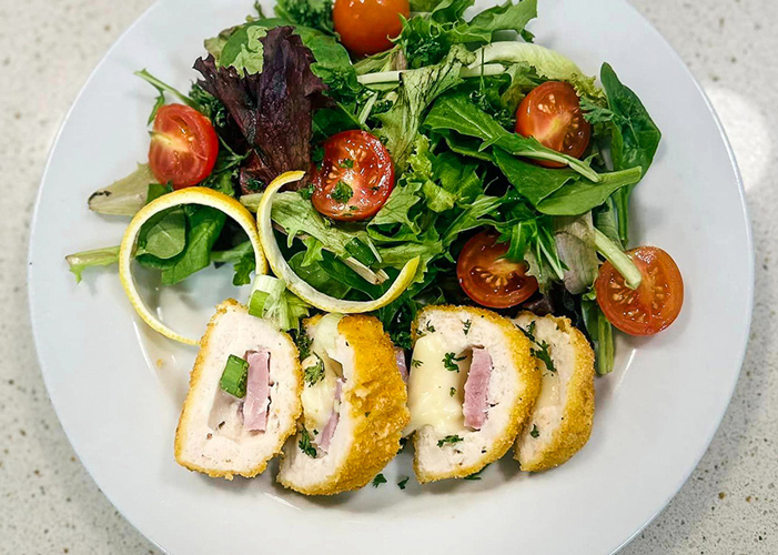 Plate featuring golden breaded chicken cordon bleu slices filled with ham and melted cheese, accompanied by a fresh green salad with cherry tomatoes and spiralized cucumber garnish.