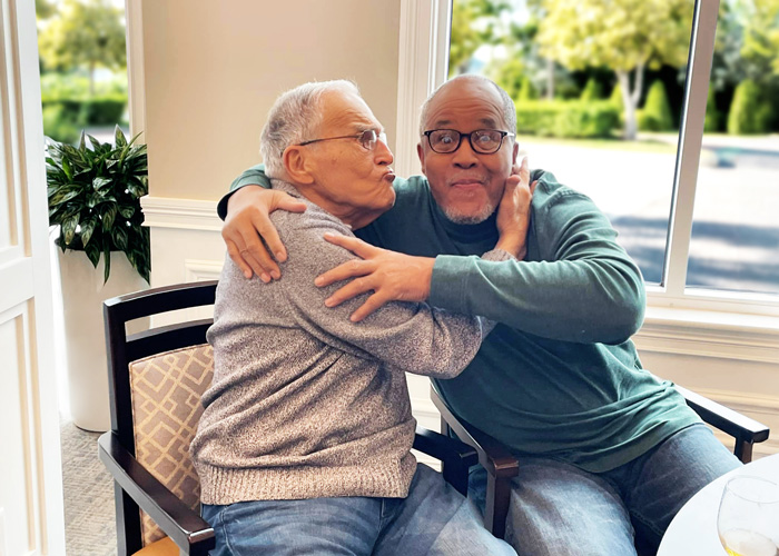 Two senior residents sharing a lighthearted moment, with one giving the other a playful kiss on the cheek while they sit together by a window, laughing and enjoying each other's company.