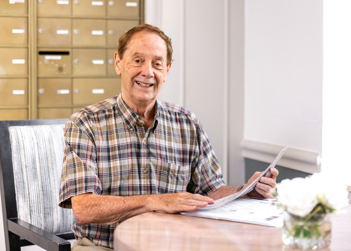 Smiling older man in a plaid shirt sits at a small round table reading a newspaper, with a background of a gold mailroom and a vase of white flowers on the table.