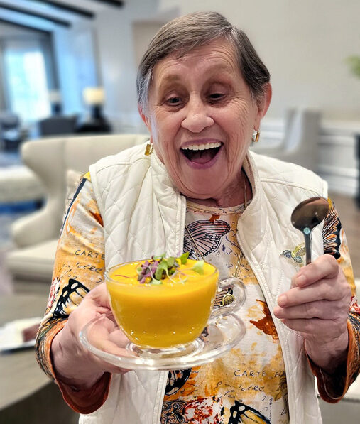 Smiling senior resident joyfully holds a bowl of vibrant orange soup garnished with greens, wearing a colorful butterfly-patterned shirt and white vest, ready to enjoy the meal.