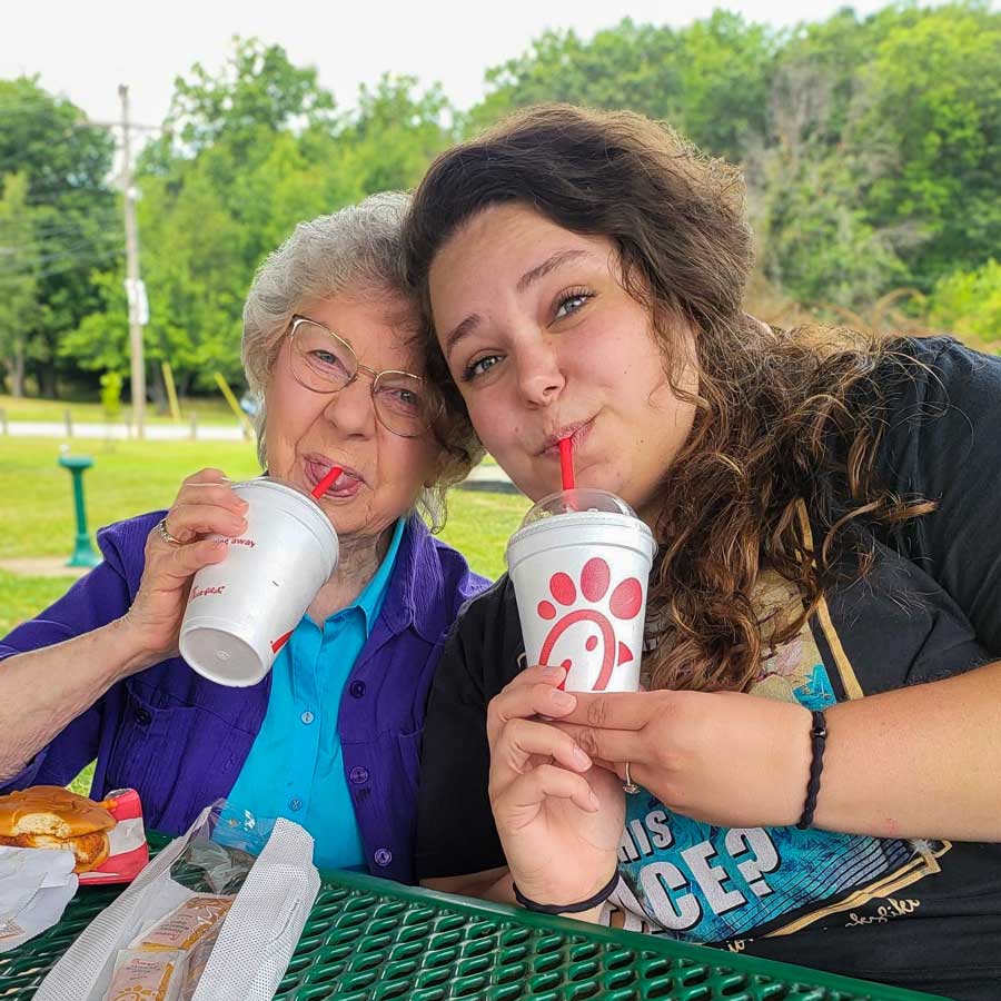 Senior resident and young woman enjoy a picnic at a park, sipping on Chick-fil-A milkshakes and smiling at the camera while sharing a meal on a green outdoor table.