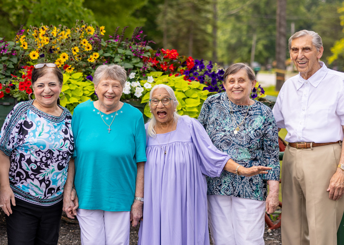 Group of five senior resident friends standing together in front of a vibrant flower display, smiling and enjoying each other's company on a beautiful day.