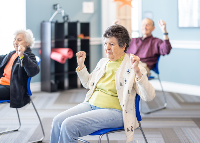 A senior resident in a light cardigan participates in a seated exercise session, raising her arms alongside other seniors in a bright room with blue chairs and exercise equipment.