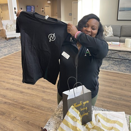 Smiling woman proudly holding up a black T-shirt with a logo, standing in a well-lit room, next to a gift bag that reads 'Thank you for being awesome' on a nearby table.