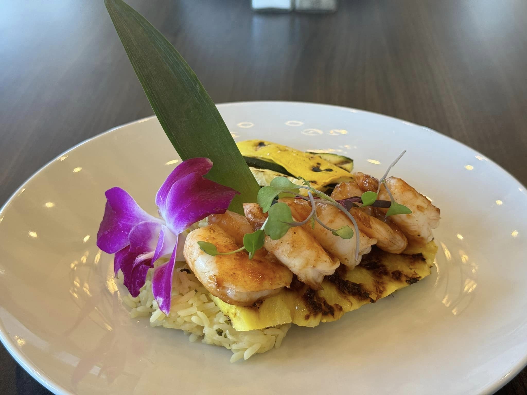 Grilled shrimp served atop a bed of rice, garnished with a purple orchid flower, pineapple slices, and microgreens, presented on a white plate with an elegant tropical flair.