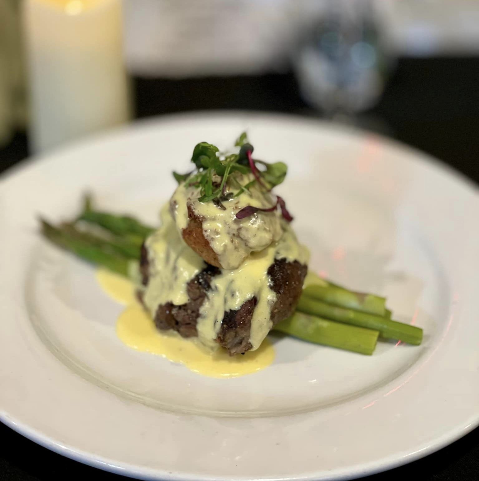 Plated gourmet steak with asparagus, topped with creamy hollandaise sauce and garnished with microgreens, set against a blurred background of a candle, flowers, and drink.