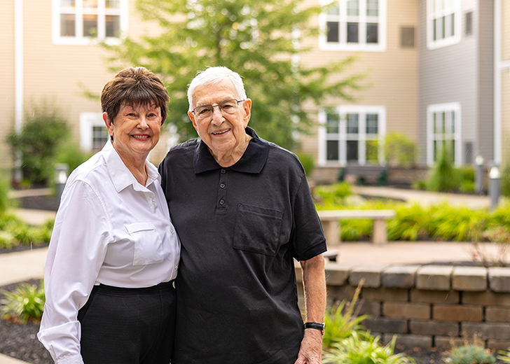 Senior resident couple standing closely together, smiling warmly at the camera, with greenery and a building in the background on a pleasant day.