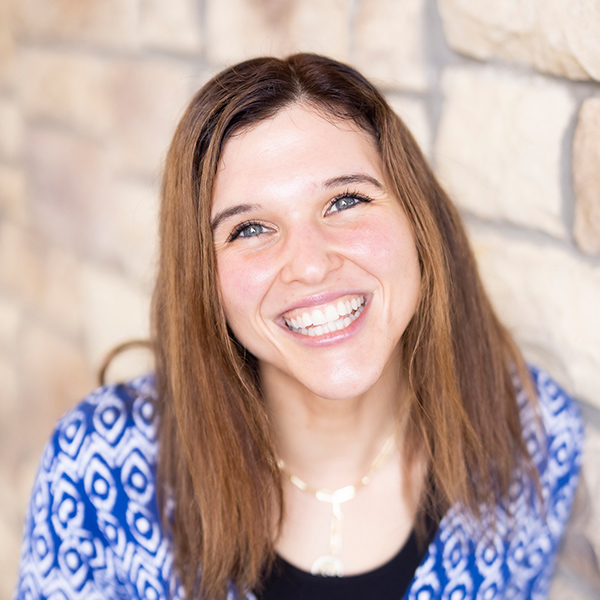 Kasey Kipping, Senior Living Director, smiling warmly in front of a stone wall, wearing a blue patterned blouse and gold necklace, radiating a friendly presence.