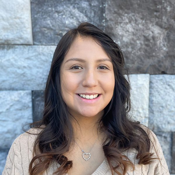 Ashley Pate, Operations Coordinator at The Barrymore Senior Living, smiling in front of a gray stone wall while wearing a light sweater and a heart-shaped necklace.