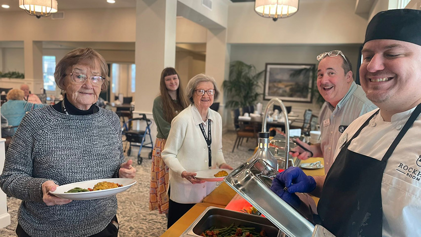 Culinary team members and residents smile near a delicious buffet in an elegant, open community space.