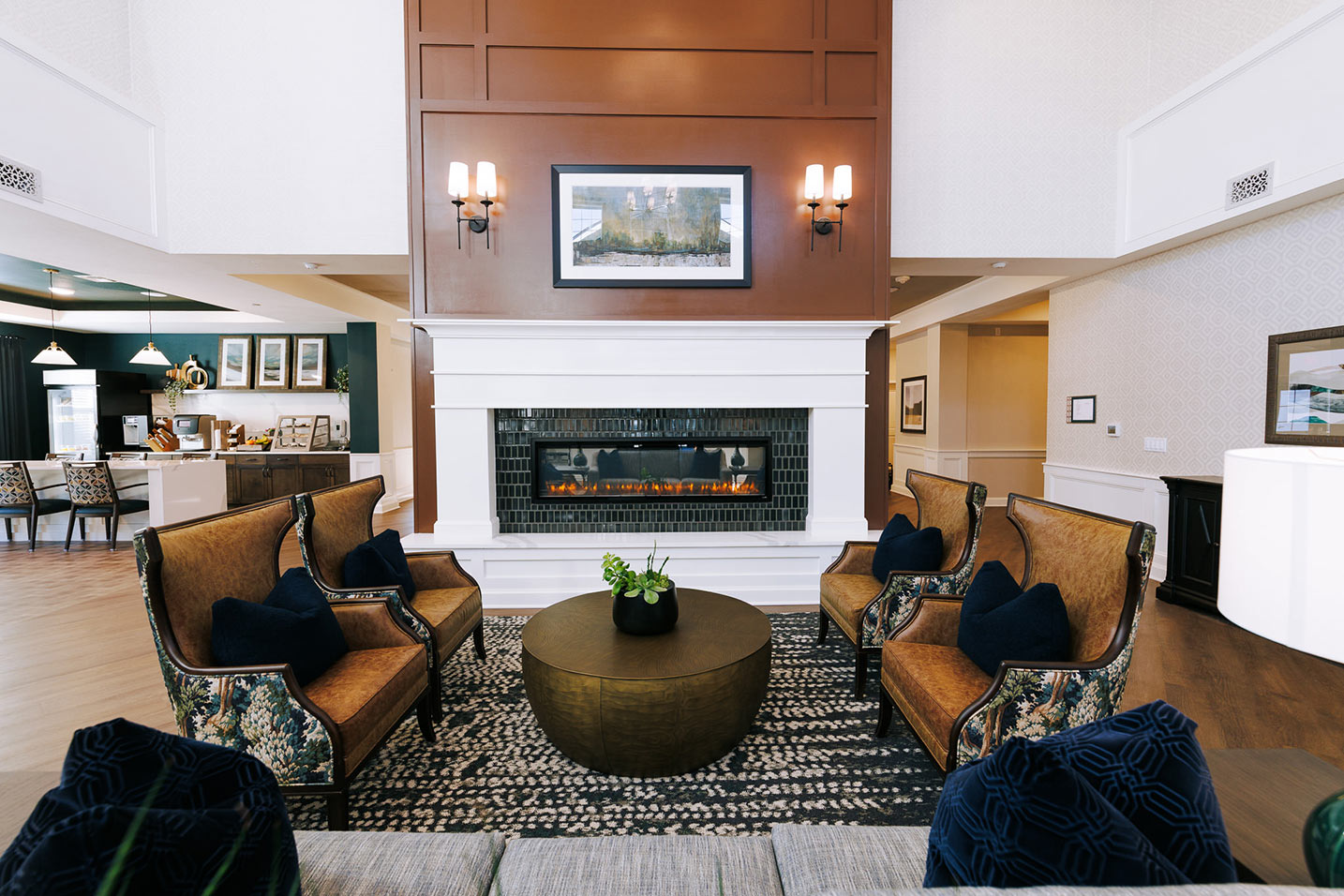 An open, elegant common space at Barrymore Senior Living, showcasing rich, inviting seating, electric fireplace, deep earth tones, and bistro area in the background.
