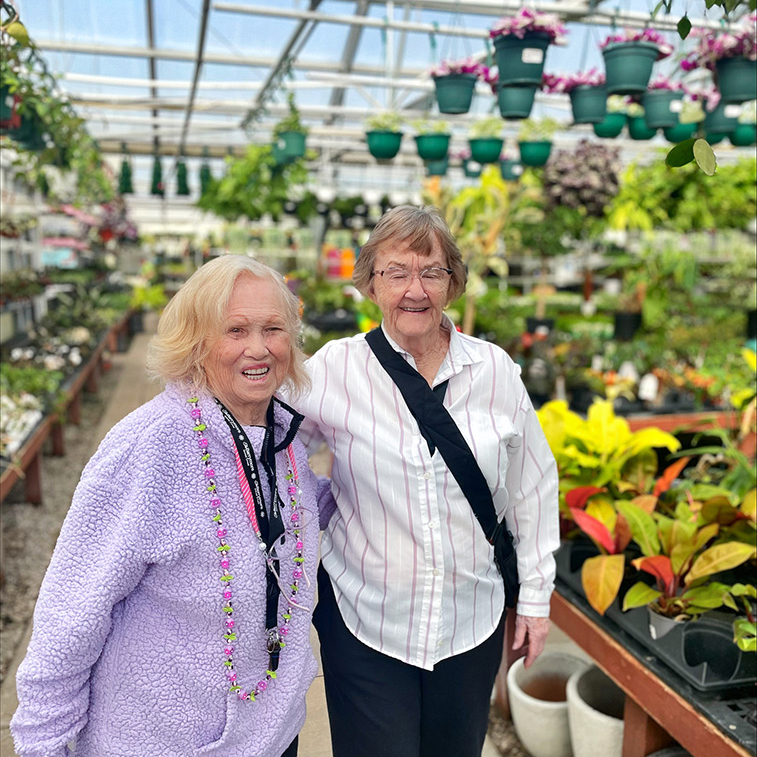 Two women smile brightly in a local greenhouse filled with plants and flowers.