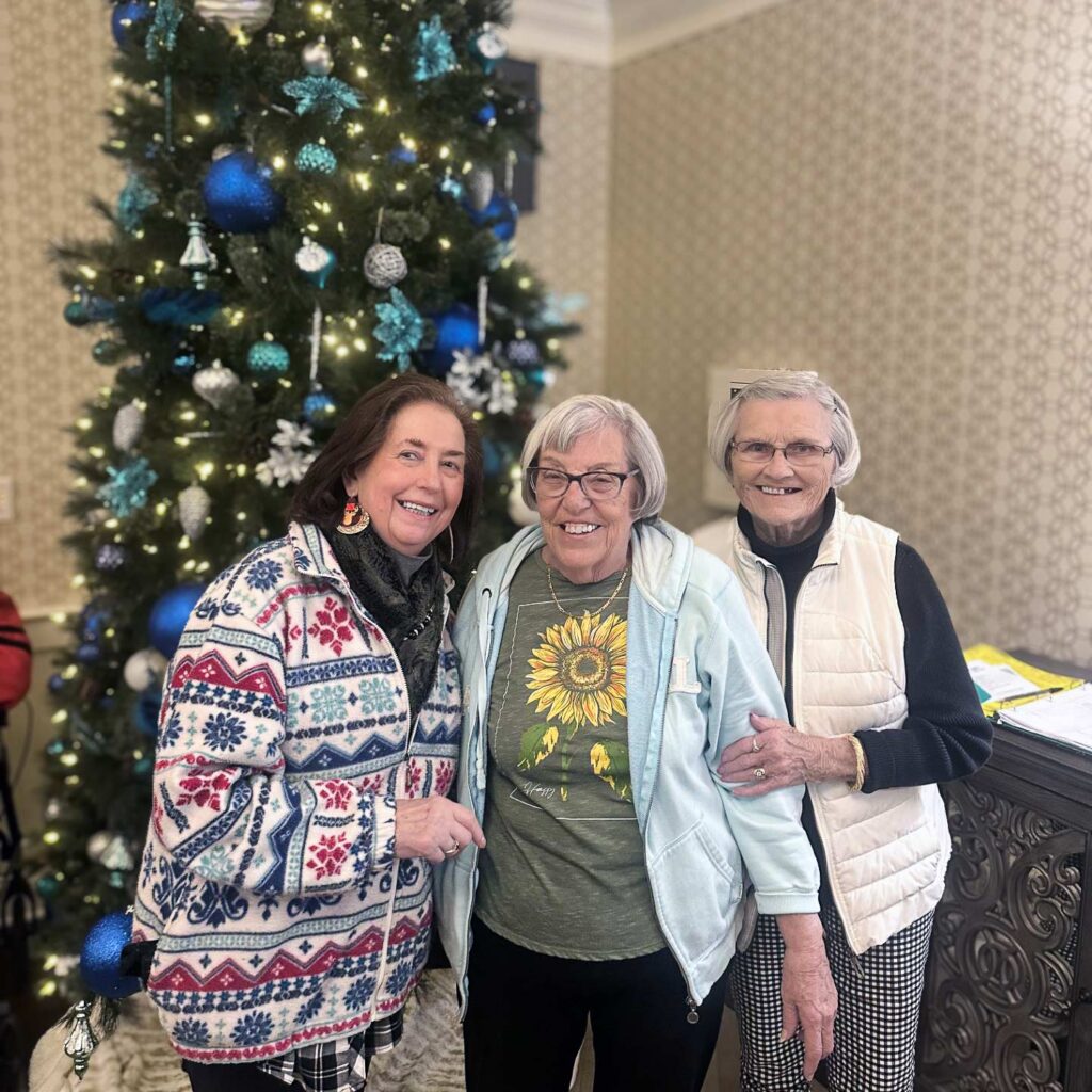 Three women smile brightly in front of a tall decorated Christmas tree at The Barrymore Senior Living.