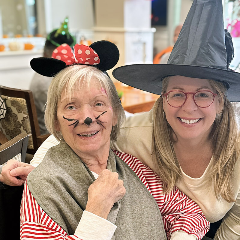 A senior woman wearing mouse ears smiles with a younger woman in a witch's hat during a community holiday event.