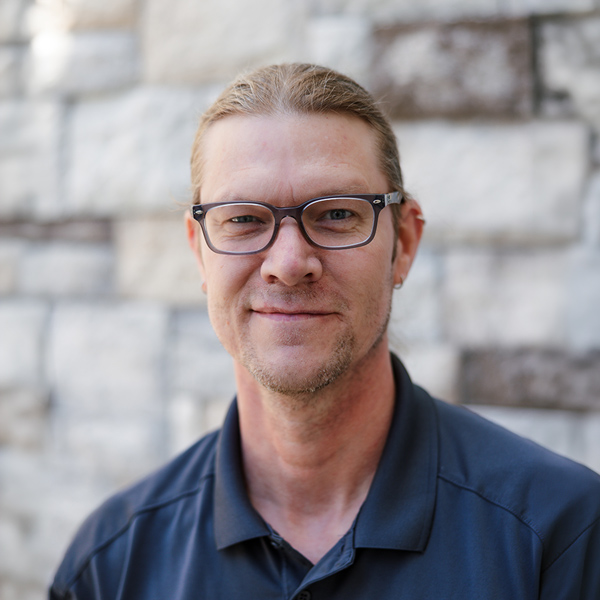 Aaron Stockwell, Plant Operations Director at The Barrymore Senior Living, looking at the camera in a professional headshot with hair pulled back, glasses, light facial hair, a dark polo shirt, and a softly blurred gray stone wall background.