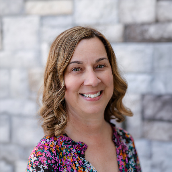 Jennifer “Jen” Craigie, Business Office Director at The Barrymore Senior Living, smiling in a professional headshot with shoulder-length wavy light brown hair, a colorful floral blouse, and a softly blurred gray stone wall background.