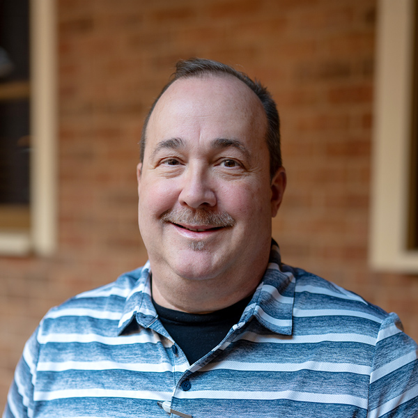 Michael Seelbach, Wellness Director at The Barrymore Senior Living, smiling in a professional outdoor headshot with short dark hair, a mustache, a blue striped polo shirt, and a softly blurred brick background.