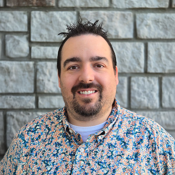 Justin Stewart, Arrow Senior Living Director providing interim support for The Barrymore Senior Living, smiling in a professional headshot with short dark spiked hair, a goatee, a colorful floral button-up shirt, and a light gray stone wall background.