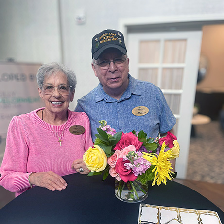 A man and woman on the resident ambassadors team at The Barrymore Senior Living smile beside a vase of fresh flowers.