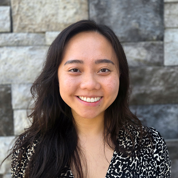 Khang Nguyen, Operations Coordinator at The Barrymore Senior Living, smiles in a professional headshot with long dark hair, a black-and-white patterned blouse, and a gray stone wall background.