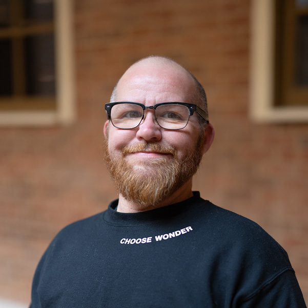 Tyler Sproston, Culinary Director at The Barrymore Senior Living, smiling in a professional headshot, wearing glasses and a black shirt, with a softly blurred brick and window background.